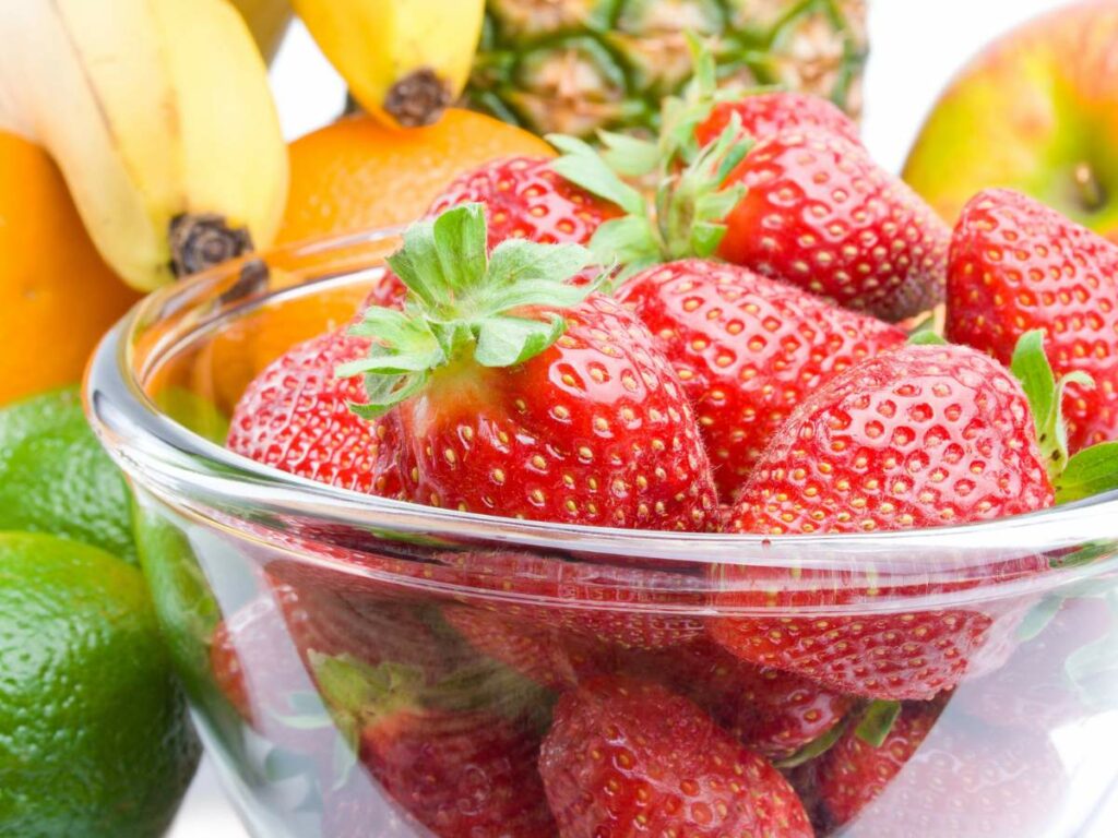 A glass bowl filled with ripe strawberries. In the background, there are bananas, oranges, a pineapple, limes, and an apple.