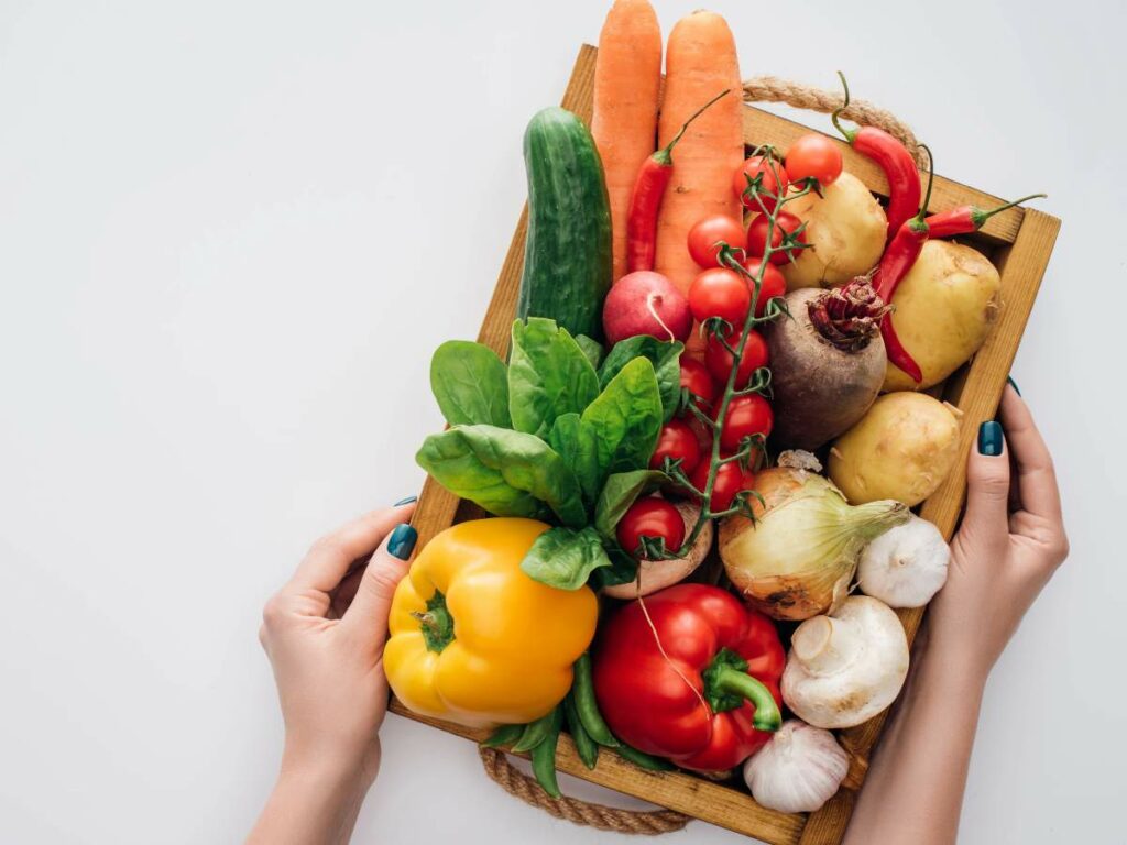 A wooden tray filled with fresh vegetables including carrots, tomatoes, peppers, onions, garlic, potatoes, a beet, a radish, spinach, and a cucumber. Two hands hold the tray.