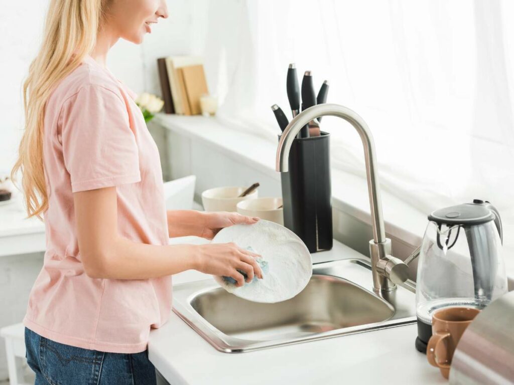 A person in a pink shirt washes dishes at a kitchen sink.