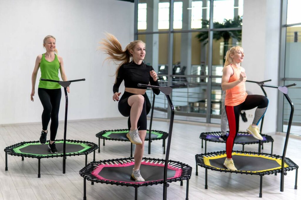 Three women are exercising on mini trampolines in a brightly lit room with large windows. They are performing a workout routine and using the trampoline handles for support.