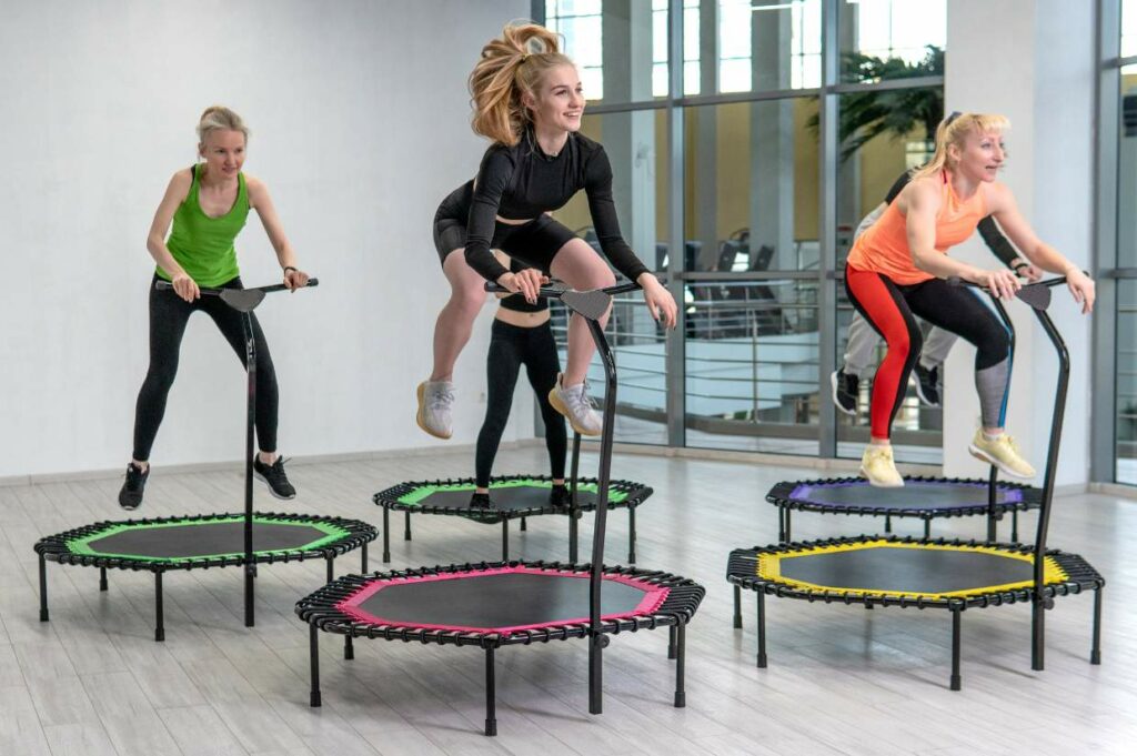 Four women are exercising on mini trampolines with handlebars in a brightly lit room. The women are mid-jump, wearing athletic clothing.