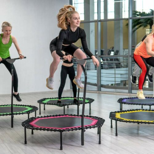 Four women are exercising on mini trampolines with handlebars in a brightly lit room.