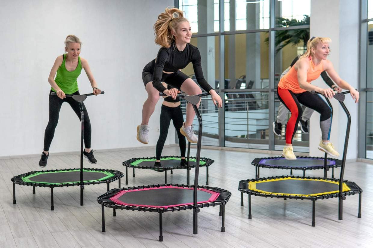 Four women are exercising on mini trampolines with handlebars in a brightly lit room.