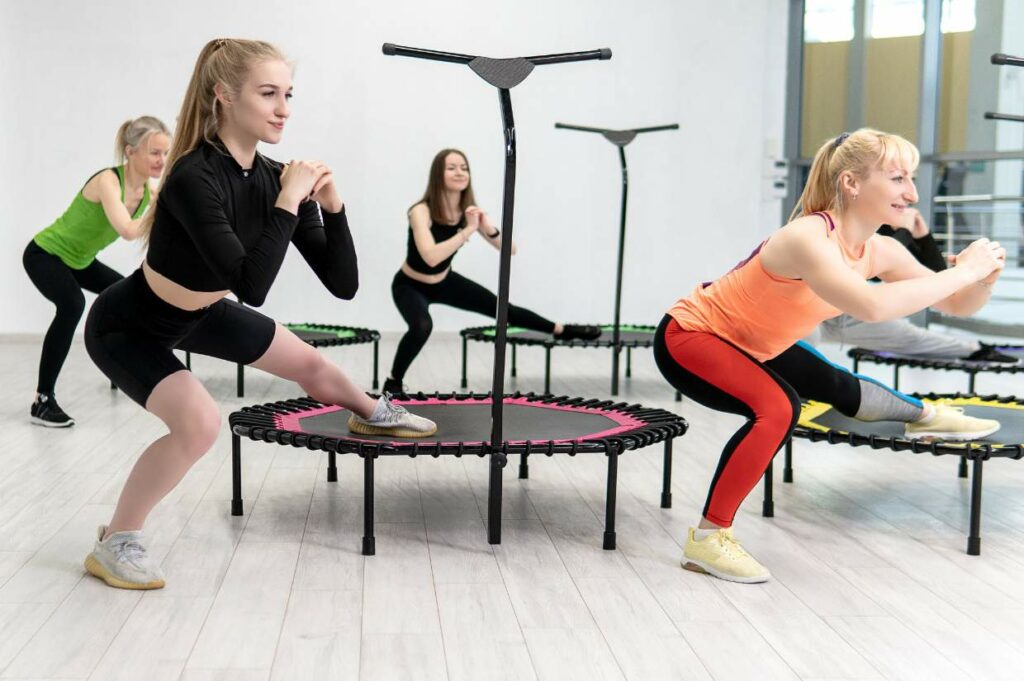 A group of women are participating in a trampoline fitness class, performing squats and stretches on mini trampolines in a brightly lit room.