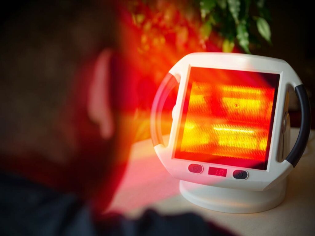 A person is sitting in front of a small, white infrared heating device with a glowing red screen.