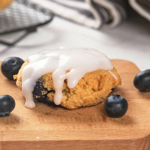 A glazed blueberry scone on a wooden board with fresh blueberries scattered around it.