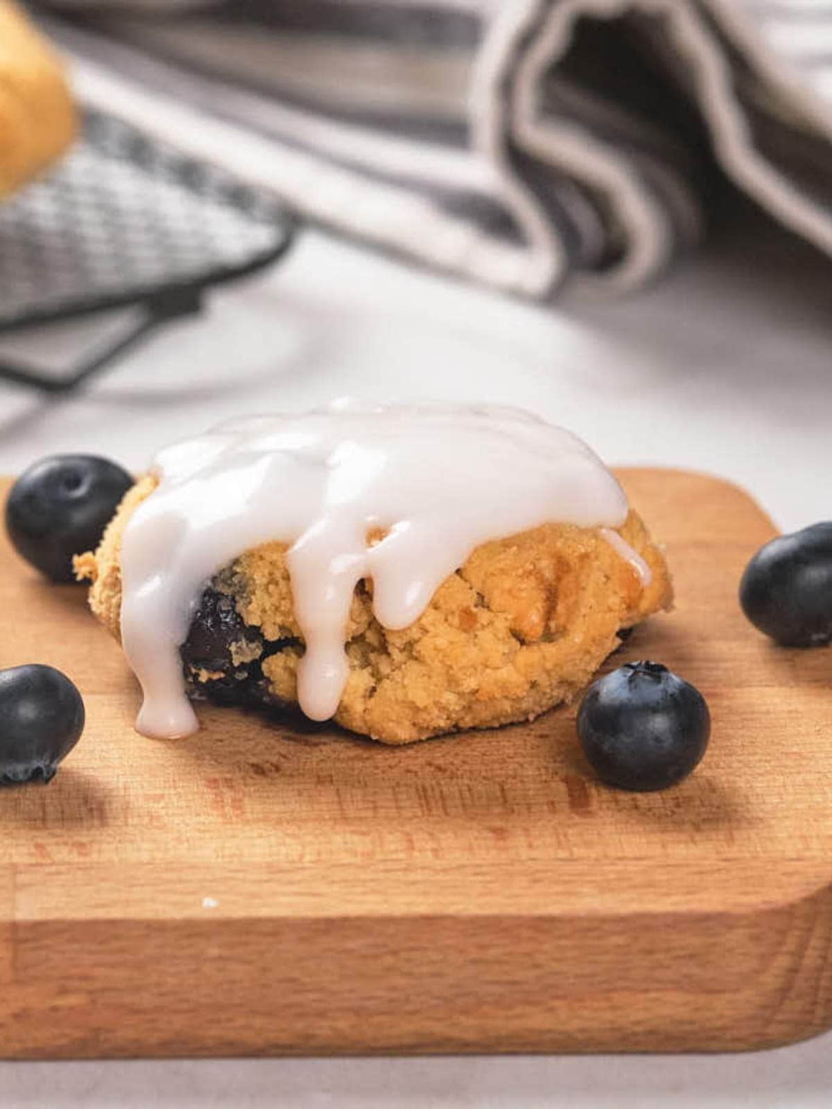 A glazed blueberry scone on a wooden board with fresh blueberries scattered around it.