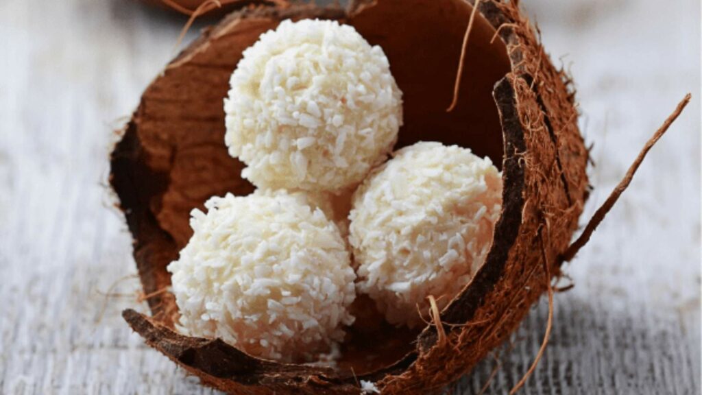 Three coconut-covered white chocolate truffles sit inside a halved coconut shell on a wooden surface.