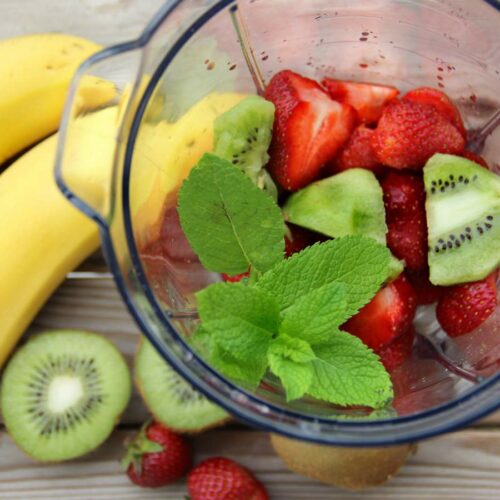 A blender with strawberries, kiwi, and mint leaves, next to two whole bananas, kiwi slices, and strawberries on a wooden surface.