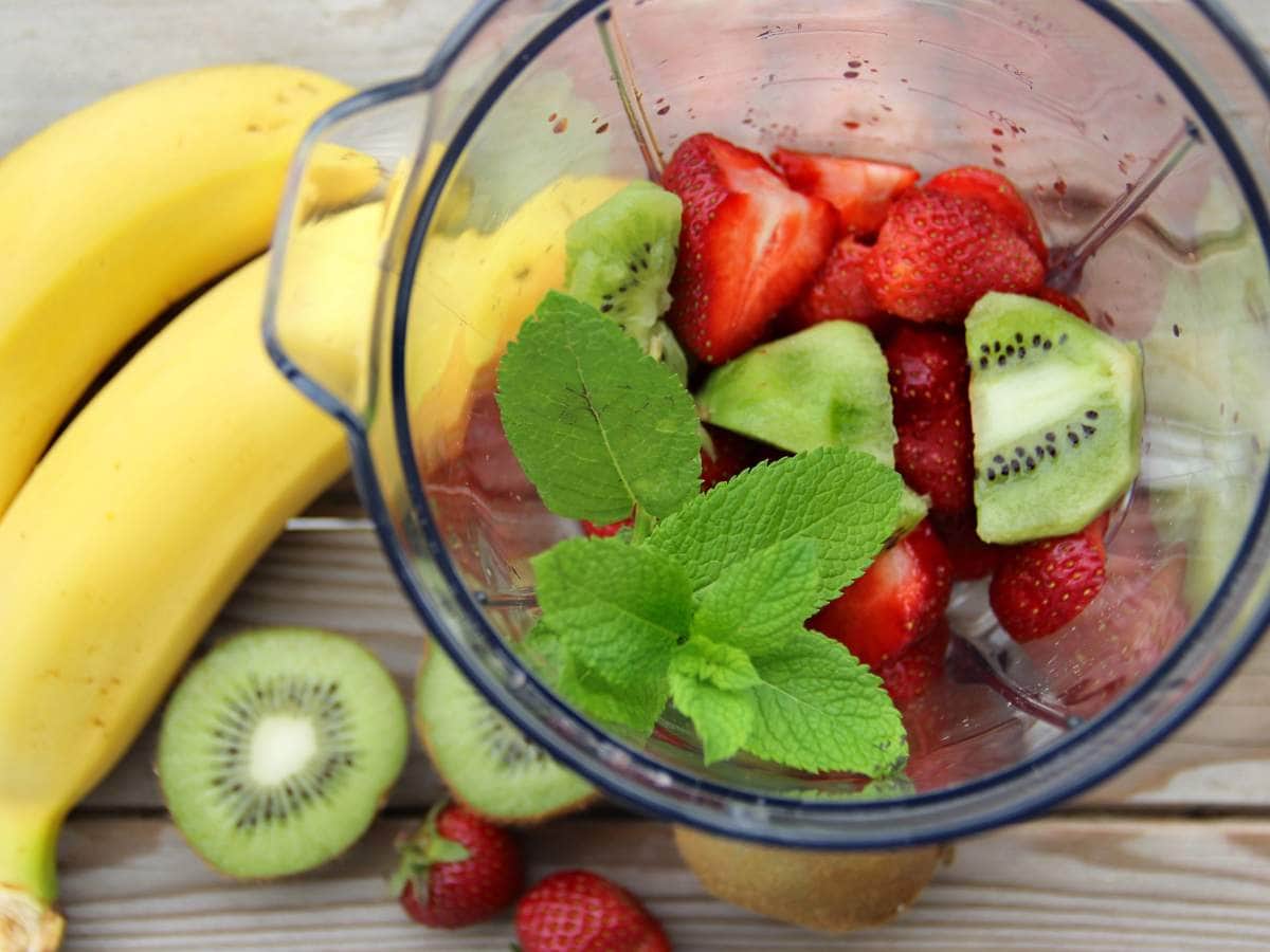 A blender with strawberries, kiwi, and mint leaves, next to two whole bananas, kiwi slices, and strawberries on a wooden surface.