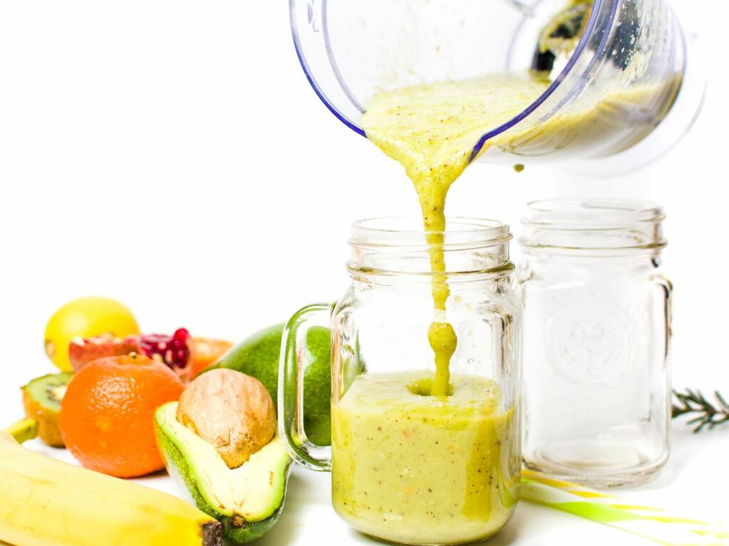 A blender is pouring a green smoothie into a glass jar, surrounded by various fruits.