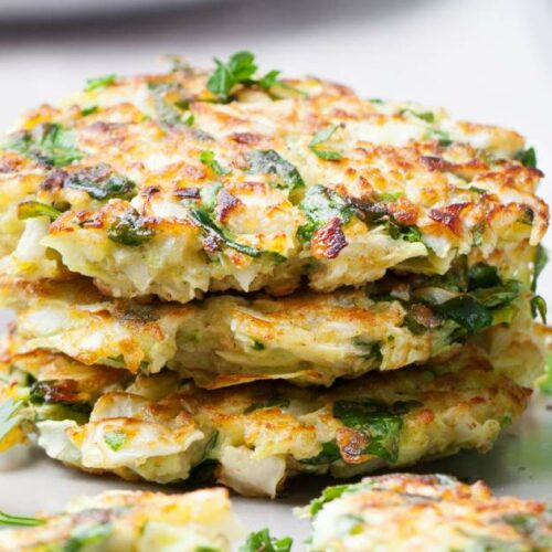 A stack of vegetable fritters garnished with herbs, placed on a plate next to a small bowl of dipping sauce.