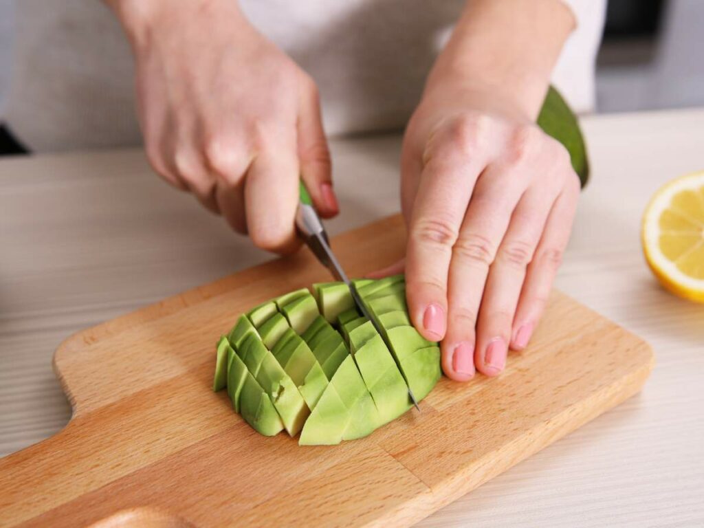 Hands chopping a peeled avocado into cubes on a wooden cutting board next to half a lemon.