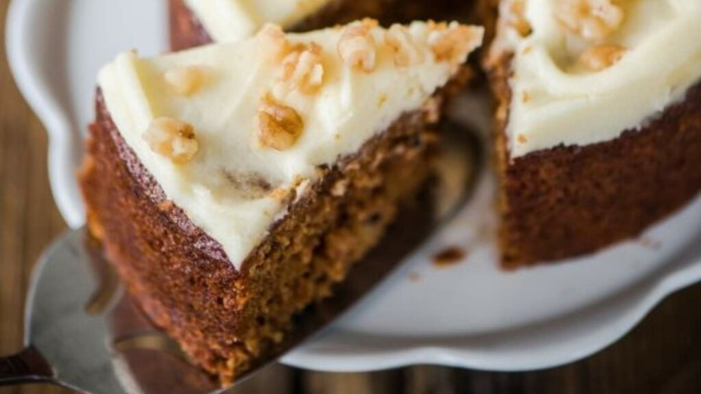 A slice of frosted carrot cake topped with nuts, being served from a larger cake.