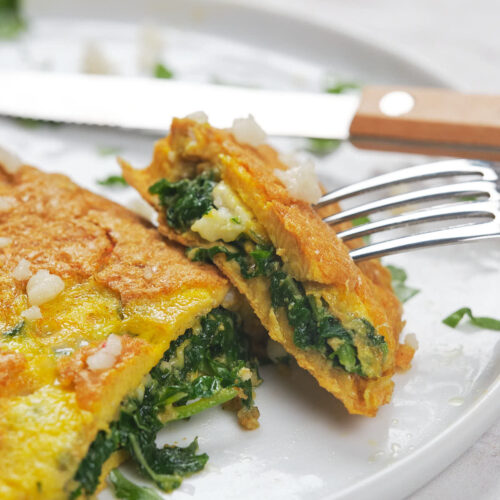 A fork and knife are used to cut into a spinach and cheese omelette on a white plate.