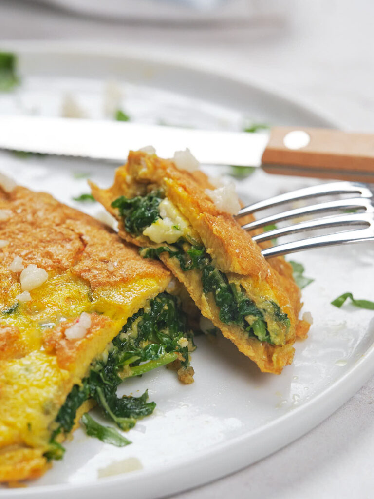 A fork and knife are used to cut into a spinach and cheese omelette on a white plate.