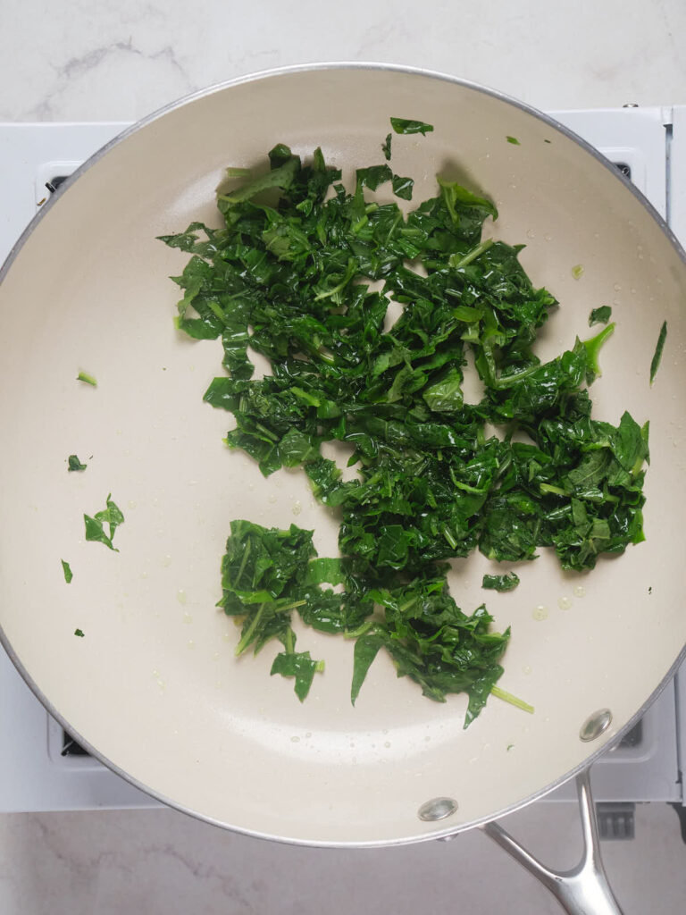 Chopped greens being sautéed in a frying pan over a stove.