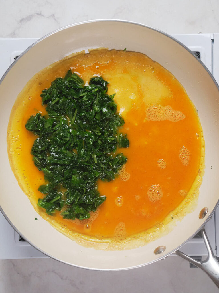 A pan on a stovetop containing a cooking omelet with bright green spinach on one side.