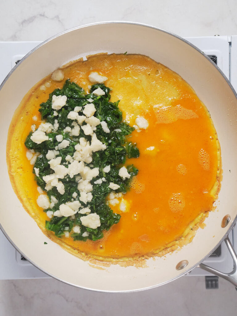 An omelette being cooked in a pan, with one side filled with chopped spinach and crumbled cheese.