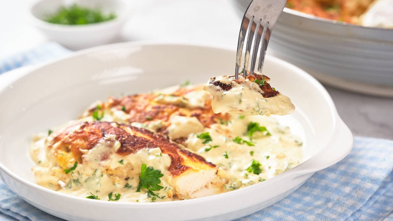 A dish with chicken breasts in a creamy white sauce garnished with chopped parsley. A fork is lifting a piece of chicken above the plate. A bowl of herbs is in the background.