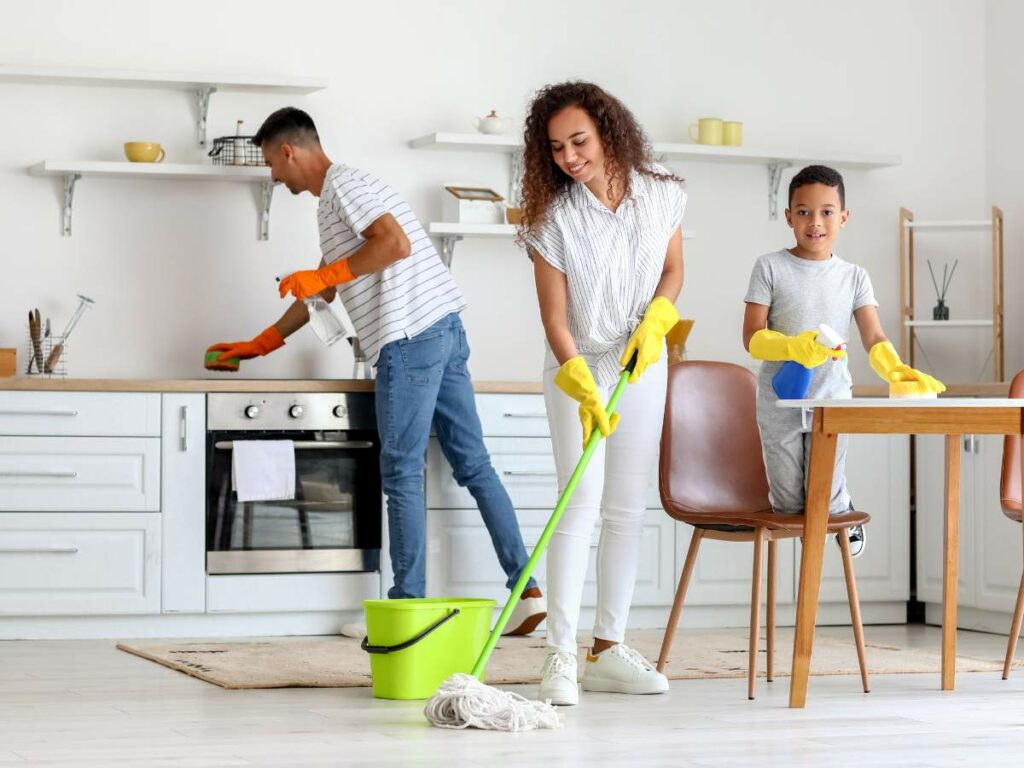 A family of three cleans a kitchen. The man is wiping the stove, the woman is mopping the floor, and the child is cleaning the table.