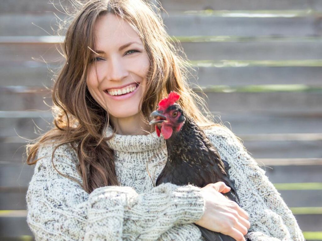 A woman smiles while holding a black chicken in front of a wooden fence.