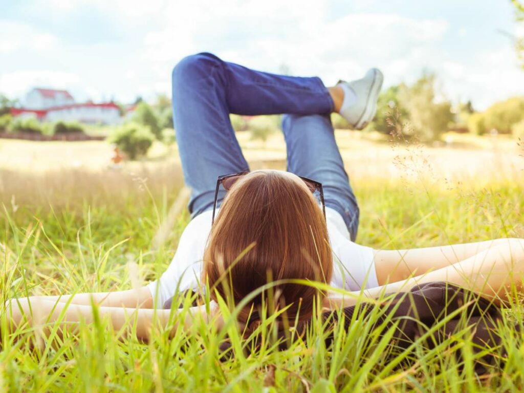 A person lies on their back in a grassy field, wearing jeans, a white shirt, and sunglasses.