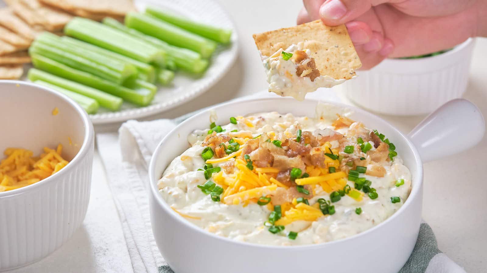 A hand dipping a cracker into a bowl of creamy dip topped with chopped green onions, bacon bits, and cheese, with a plate of celery sticks and another bowl of shredded cheese in the background.