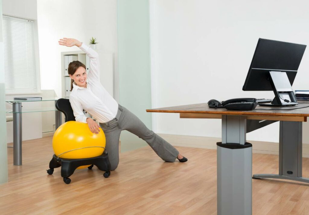 Person stretching while seated on a yellow exercise ball at an office desk, with a computer monitor on the desk.