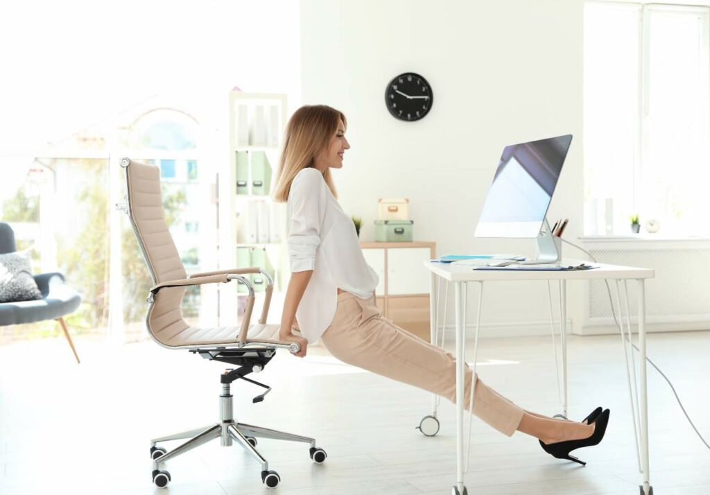 A woman in business attire stretches her legs while sitting at an office desk, smiling at a computer screen in a bright room.