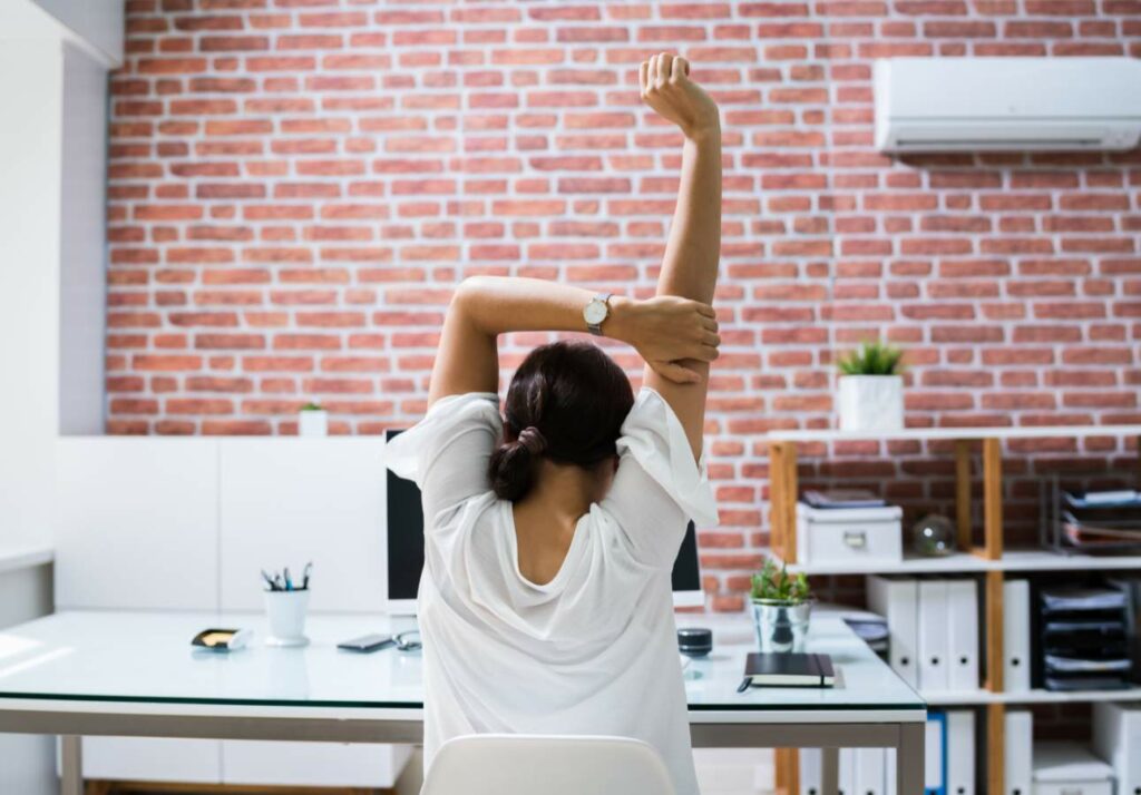 A person sitting at a desk in a modern office stretches their arm up while facing a brick wall.