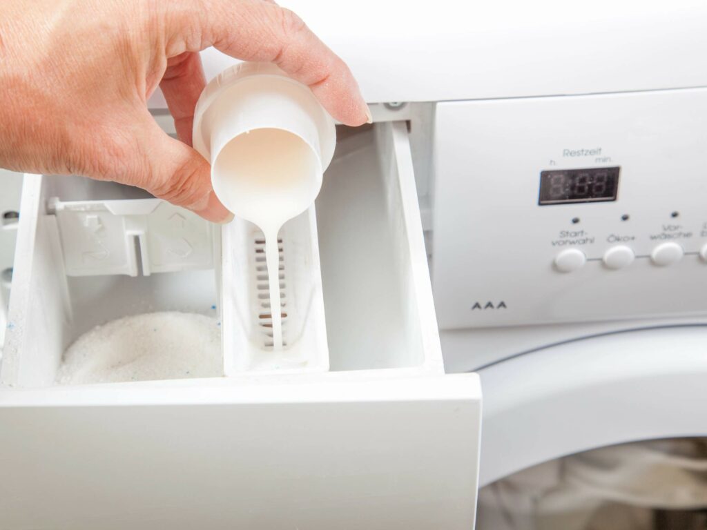 A hand pours liquid detergent into the drawer of a washing machine.