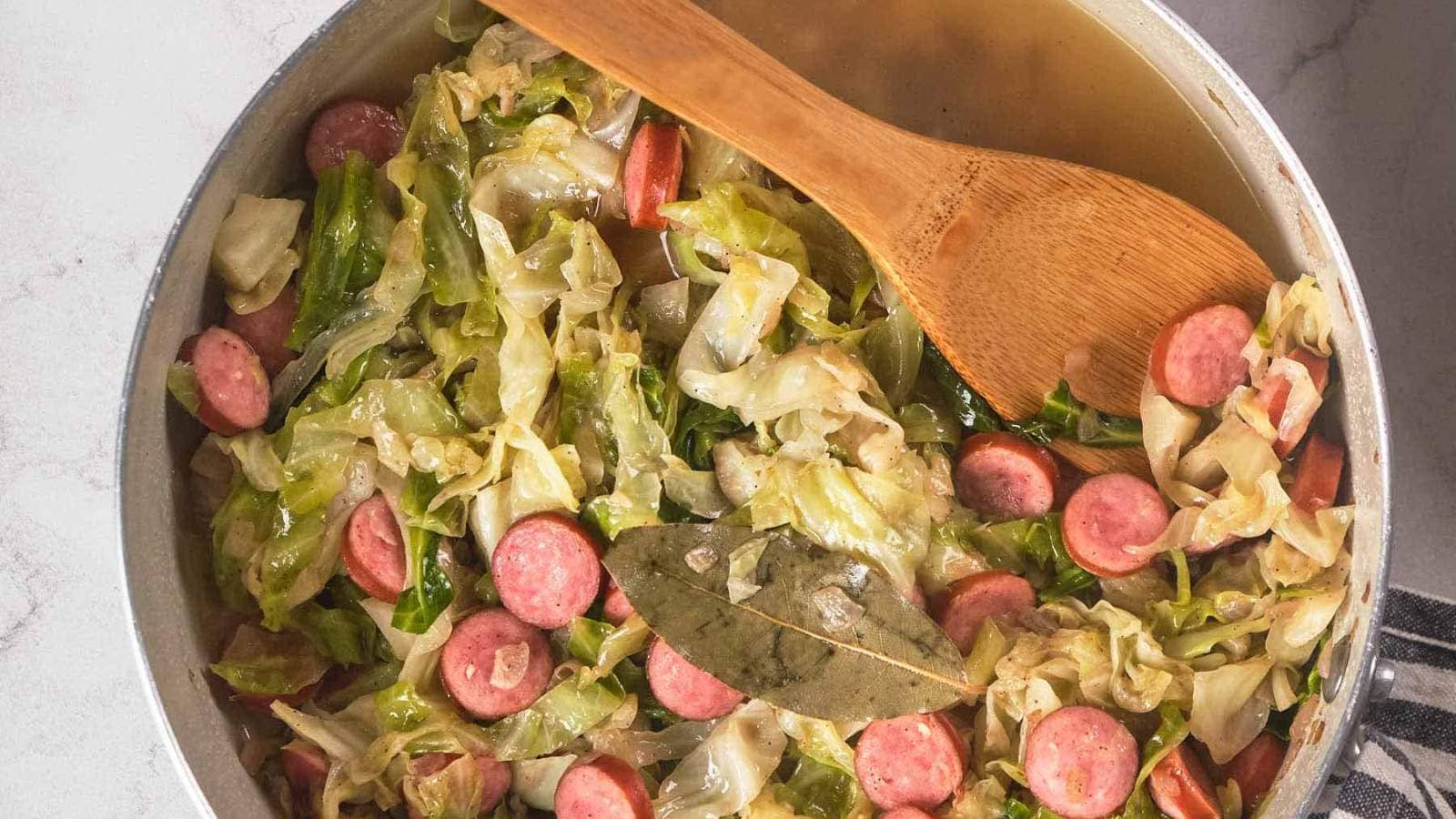 A pan filled with fried cabbage and sausage, with a wooden spoon resting inside.