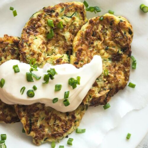 A plate of zucchini fritters topped with a dollop of sour cream and sprinkled with chopped green onions.