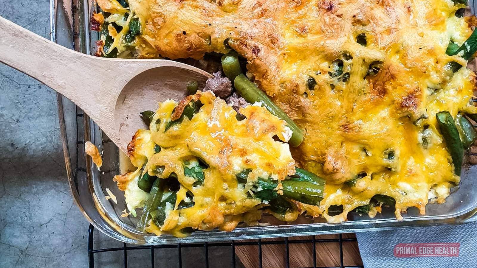 A close-up of a baked casserole with melted cheese, green beans, and vegetables being served from a glass dish with a wooden spoon.