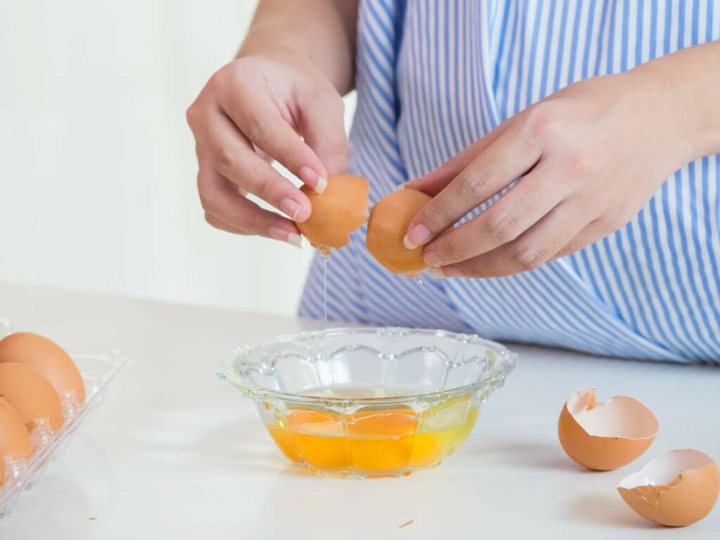 Person cracking an egg over a glass bowl with cracked eggshells and a carton of eggs nearby.