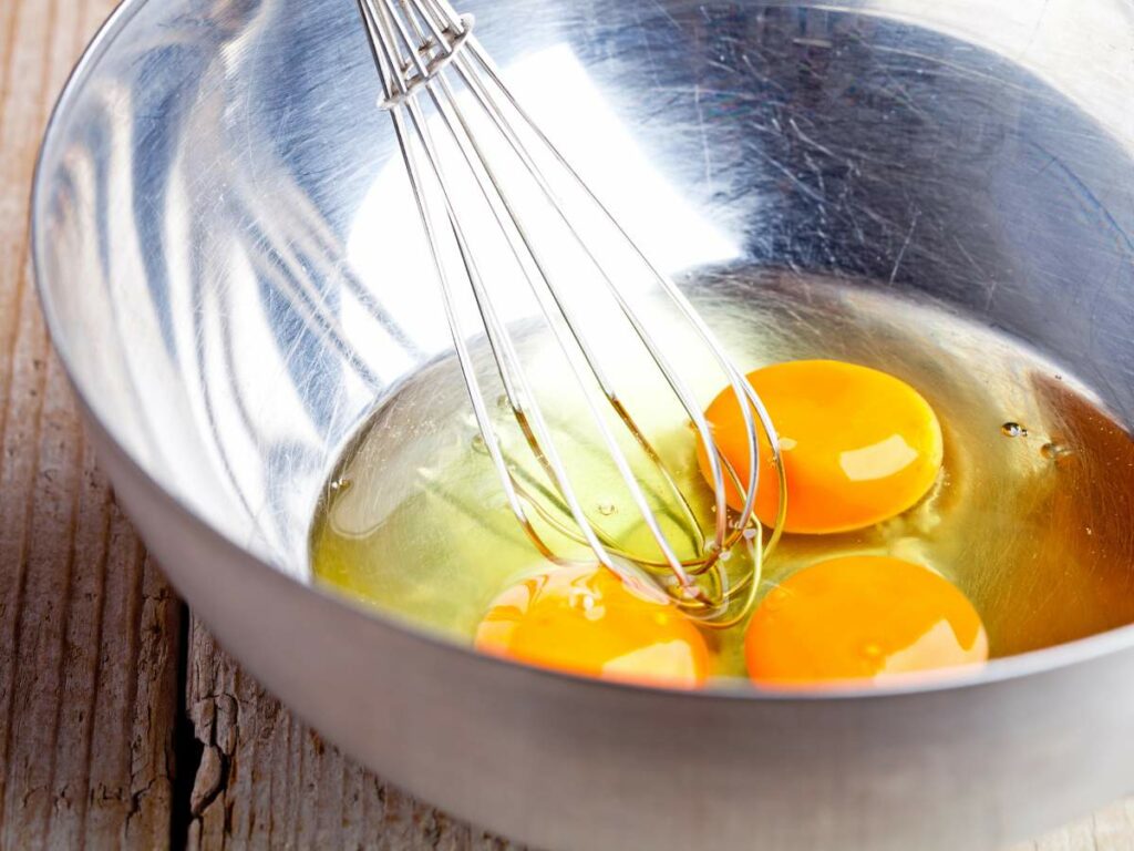 A whisk in a metal bowl contains three cracked eggs with visible yolks and whites.