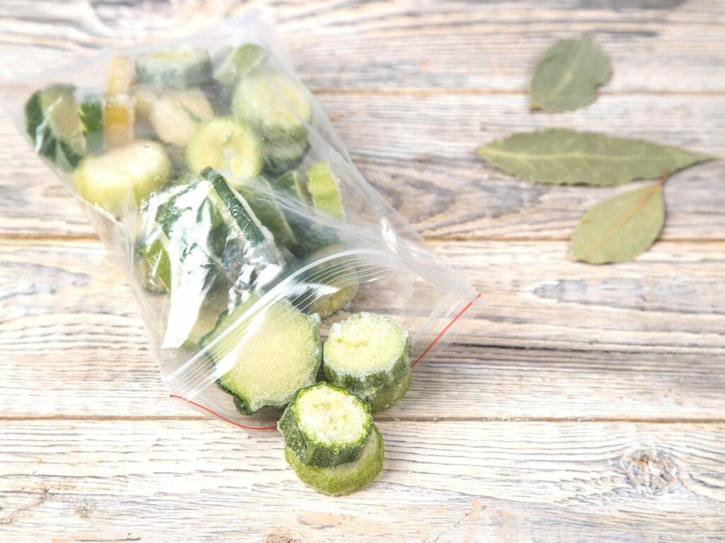 A plastic bag containing round slices of frozen zucchini on a wooden surface, with two bay leaves nearby.