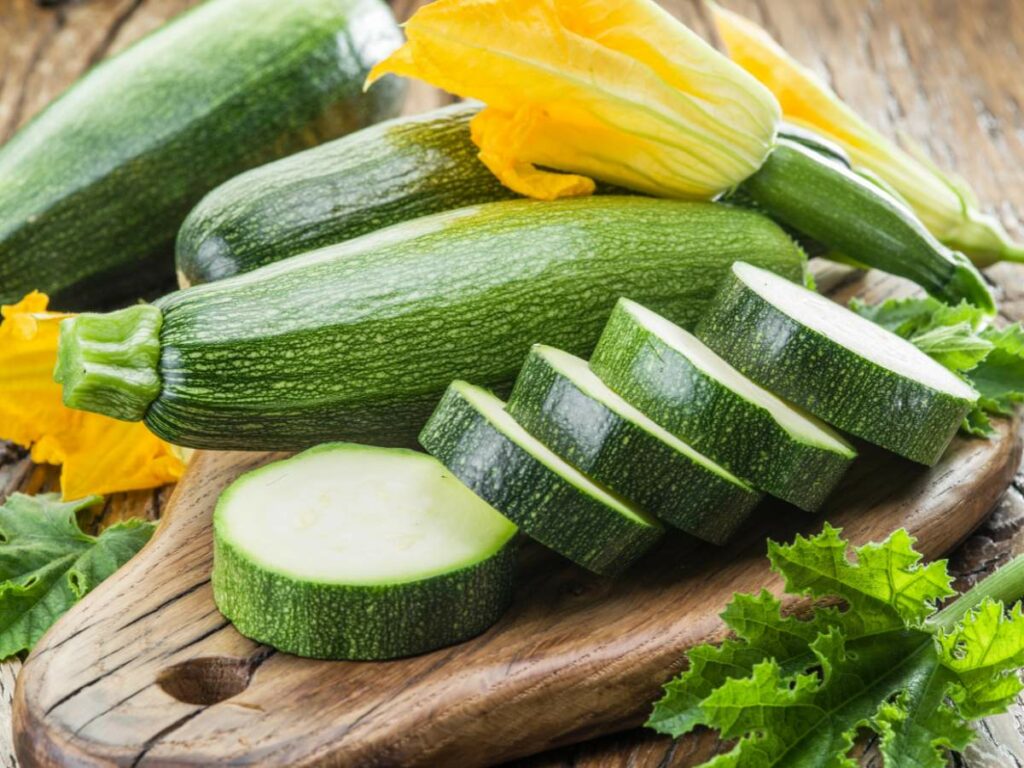 Fresh zucchinis, one with a yellow flower, and zucchini slices arranged on a wooden board with green leaves scattered around.