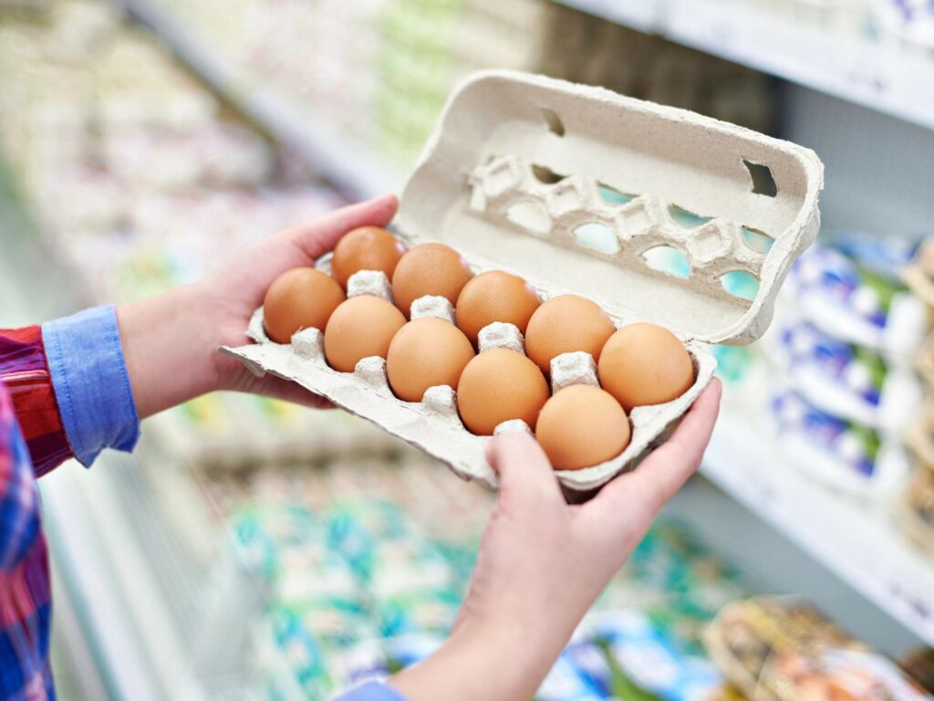 A person holds a carton of twelve brown eggs in a grocery store aisle filled with various products.