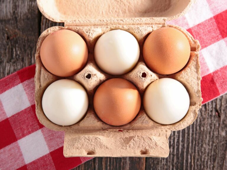 A carton containing six eggs, three brown and three white, placed on a red and white checkered cloth, resting on a wooden surface.