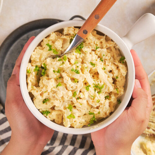 A person holds a bowl of pasta salad topped with chopped parsley.