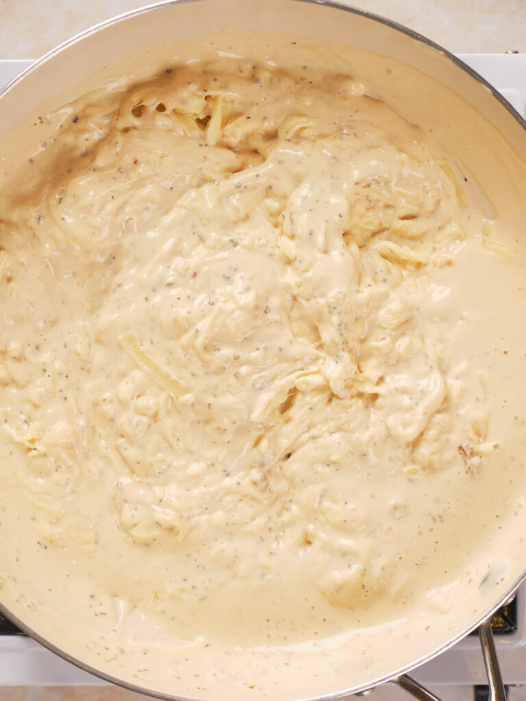 A close-up of a pan filled with creamy, light beige cheese sauce, with visible herbs and spices.