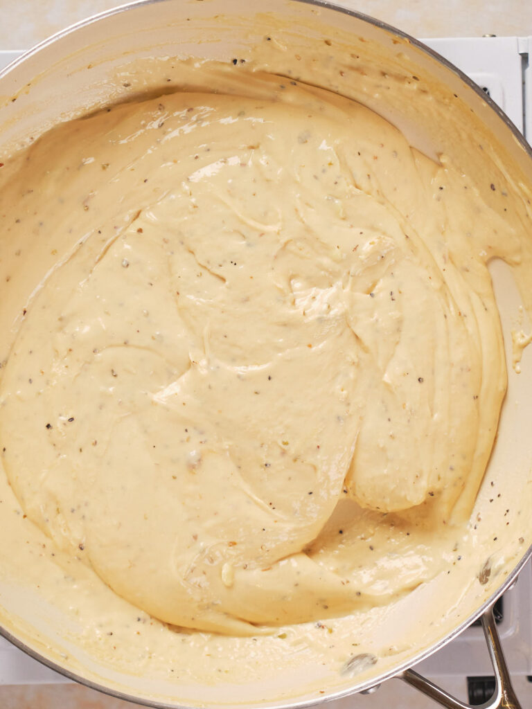 A close-up of creamy yellow cheese sauce with visible herbs cooked in a large pan.