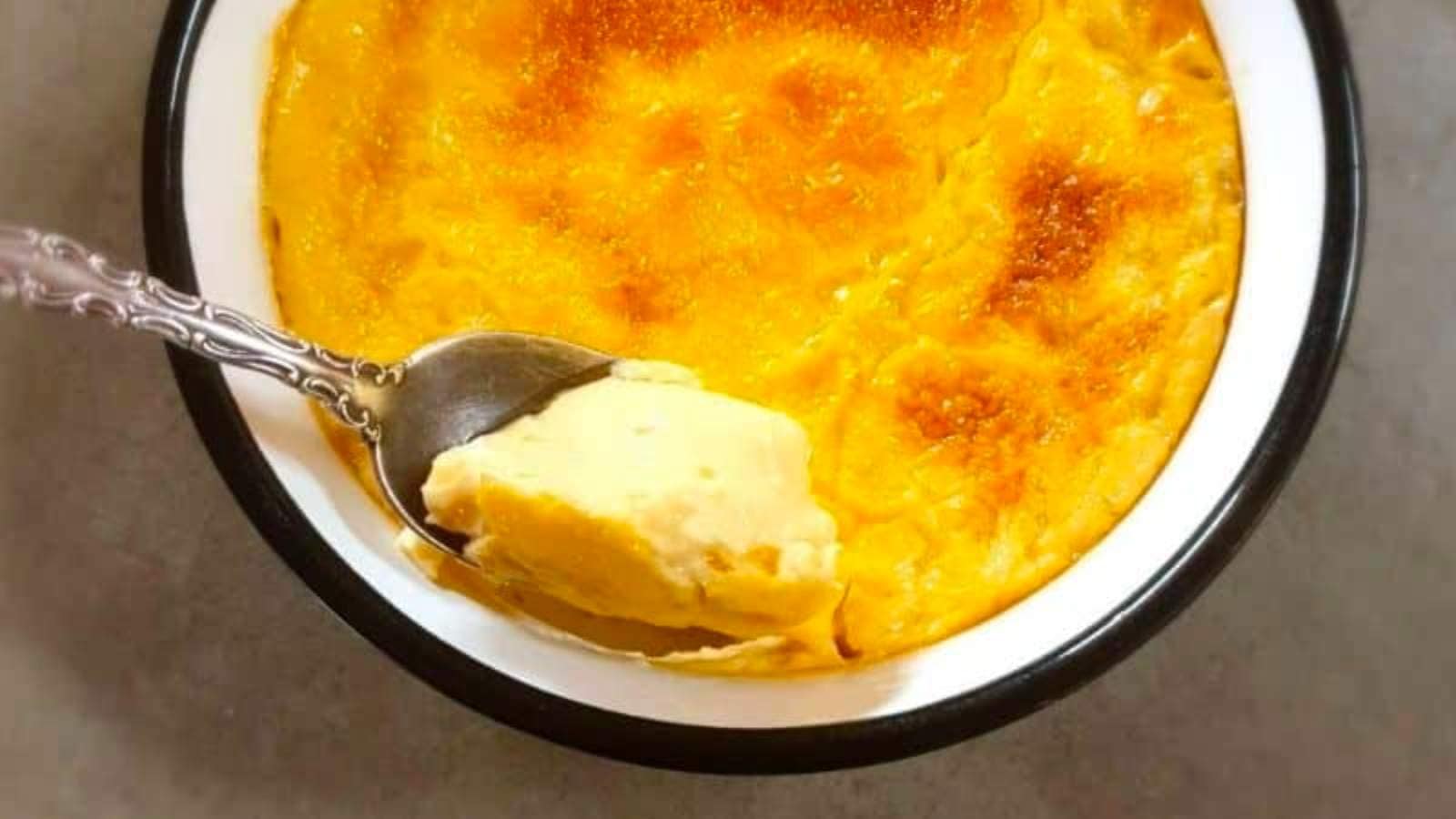 A close-up of a baked custard dessert in a round dish. A spoon has scooped out a portion, revealing its creamy texture.