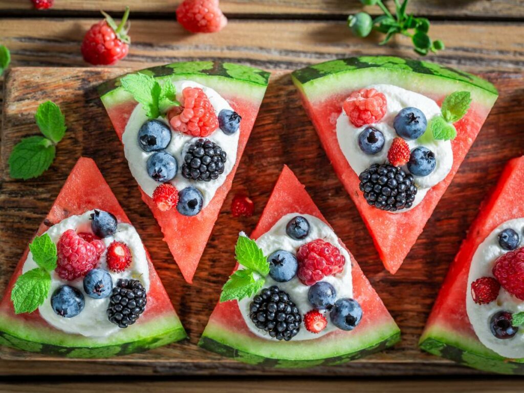 Sliced watermelon topped with whipped cream, berries, and mint leaves on a wooden surface.