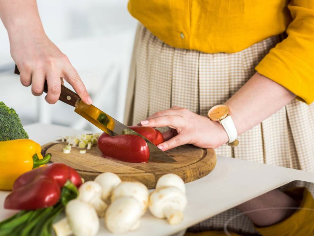 Two people preparing food; one is chopping a red bell pepper on a wooden cutting board with a knife.