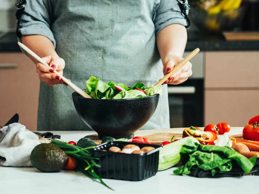 Person in apron tossing a salad with wooden spoons in a kitchen, surrounded by various fresh vegetables.