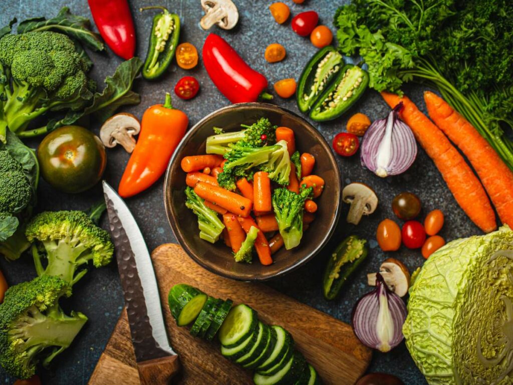 A bowl of steamed vegetables is surrounded by various fresh vegetables.