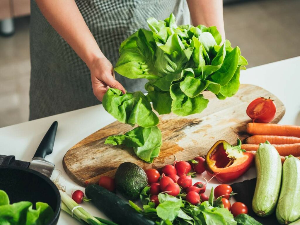 Person holding leafy greens over a wooden cutting board with assorted vegetables.